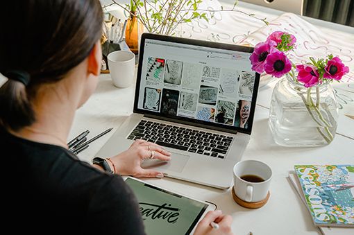 Diseñadora tomando inspiración en Pinterest, bocetando un logotipo para hacer un diseño web profesional. Ella está en una mesa, usando su laptop, tomando un delicioso café.