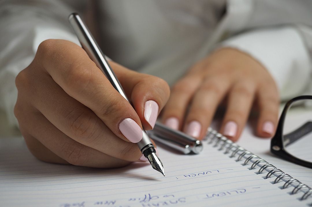 Redacción, edición, corrección de estilos. Muestra una mujer haciendo anotaciones en un cuaderno con una pluma.