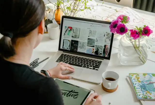 Diseñadora-inspirándose-para-diseñar-un-logotipo Diseñadora tomando inspiración en Pinterest, bocetando un logotipo para hacer un diseño web profesional. Ella está en una mesa, usando su laptop, tomando un delicioso café.
